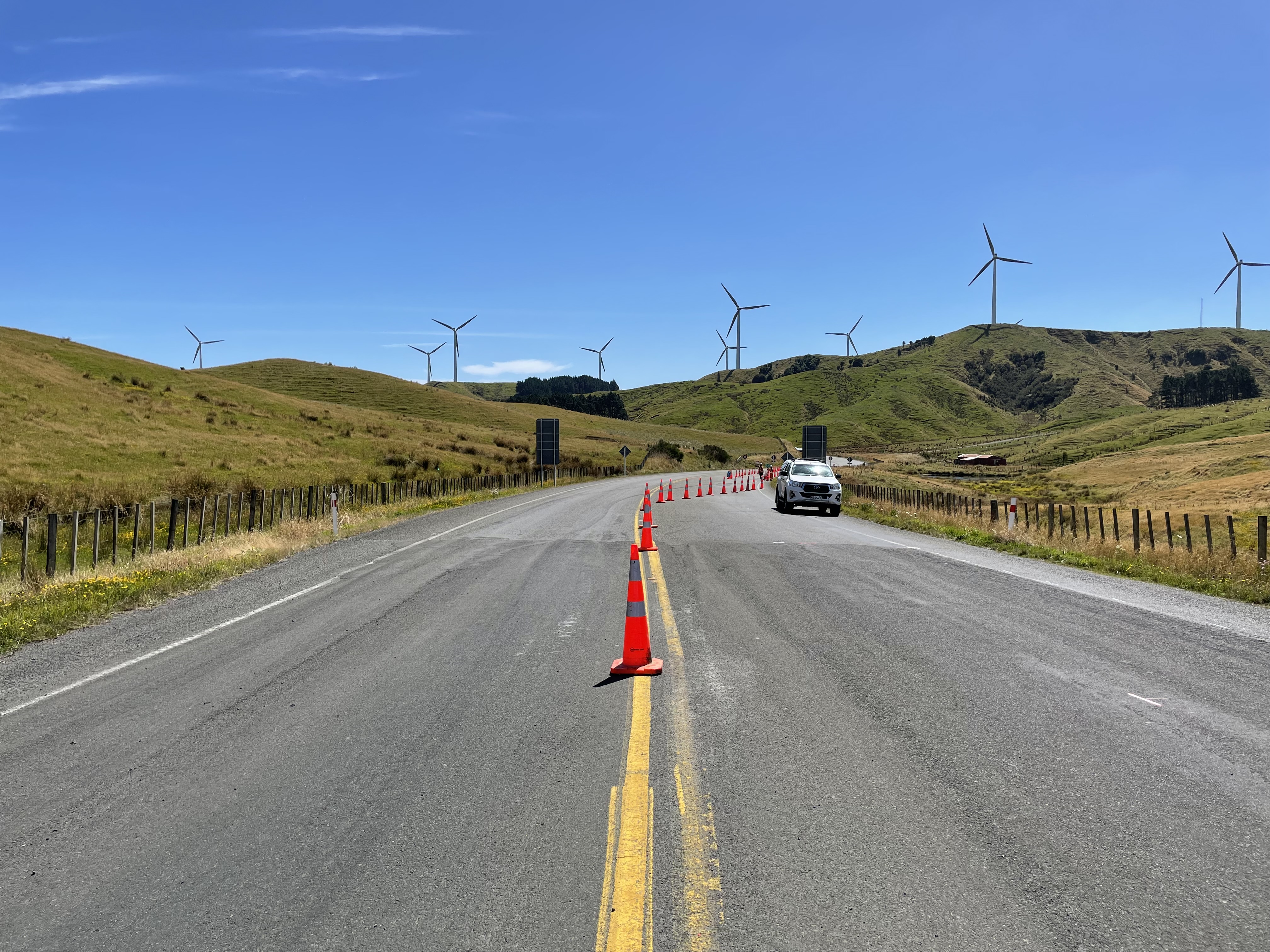 Rural road with traffic cones narrowing lanes and a white vehicle on the right side, wind turbines on grassy hills under blue sky.