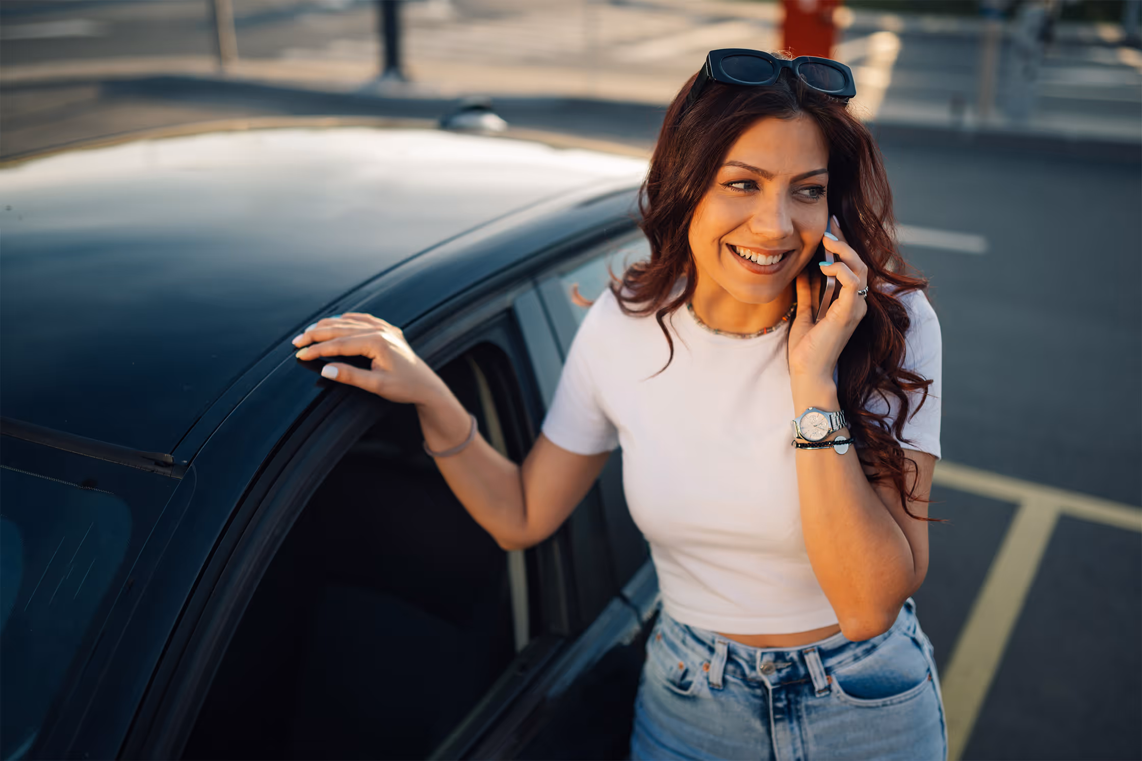 Smiling woman with sunglasses on her head talking on a cellphone while leaning on a black car door in a parking lot.