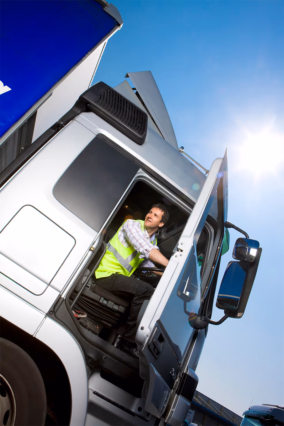 Truck driver wearing a reflective vest sitting in the cab of a white truck with the door open under a bright blue sky.