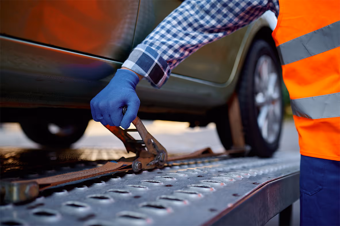 Tow truck driver wearing a checkered shirt, blue gloves, and an orange safety vest securing a strap around a car wheel on a metal flatbed.