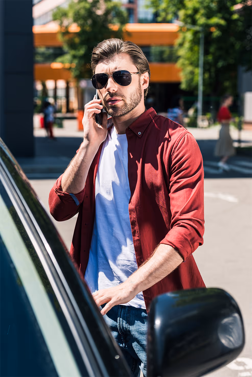 Man wearing sunglasses and a red shirt talking on a smartphone next to a car in an outdoor urban area.