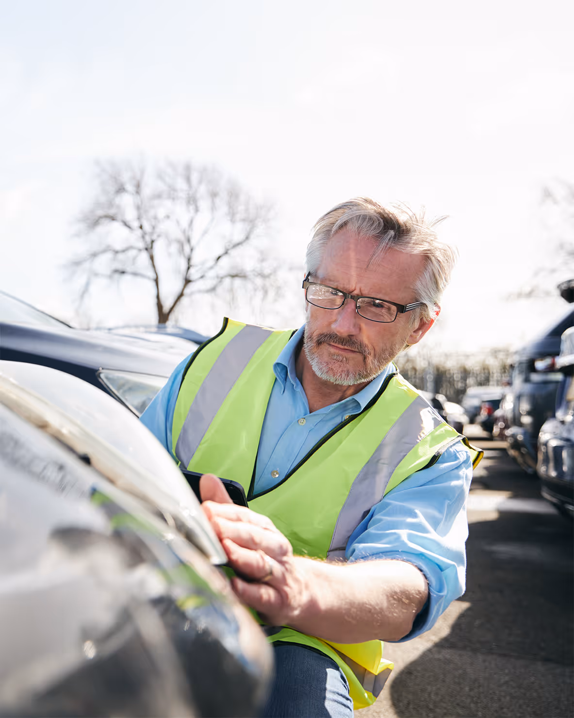 Mature man in safety vest inspecting damage on a car in a parking lot.