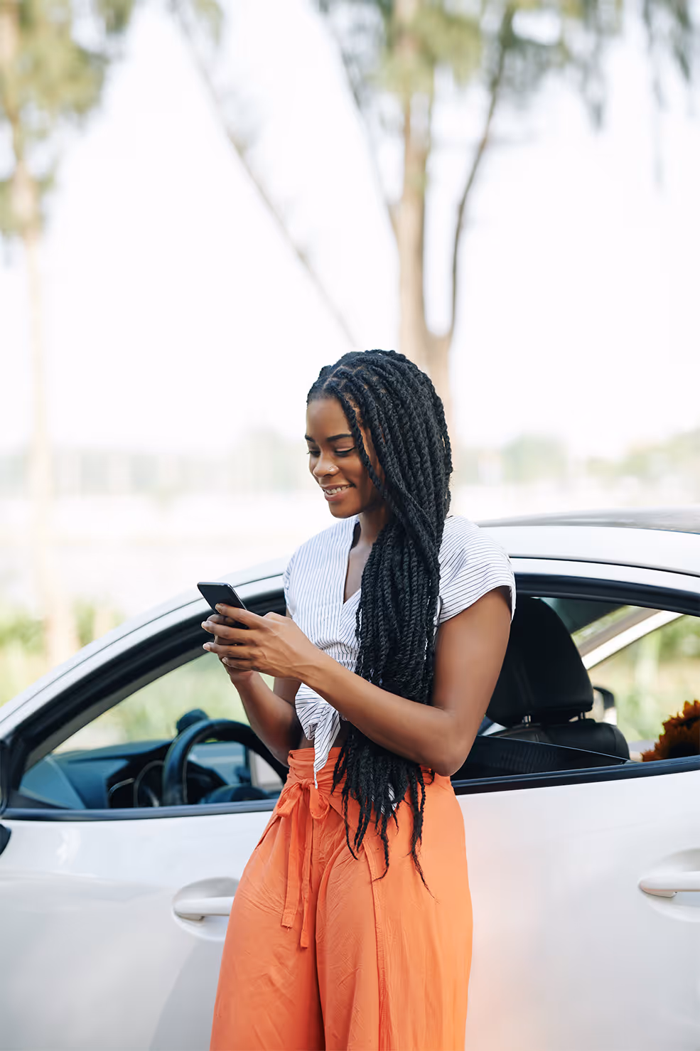 Smiling woman with long braids standing next to a white car and looking at her smartphone.
