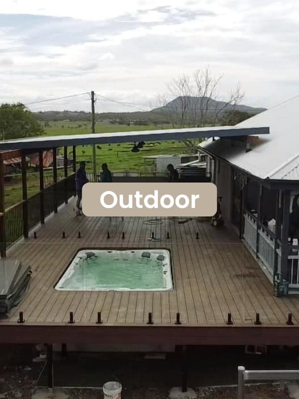 Wooden outdoor deck with a built-in hot tub and people walking under a covered area, with green fields and hills in the background.
