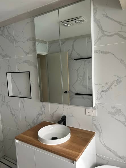 Modern bathroom with white marble walls, a wooden countertop with a round white vessel sink, black faucet, mirrored cabinet, and black towel bars.