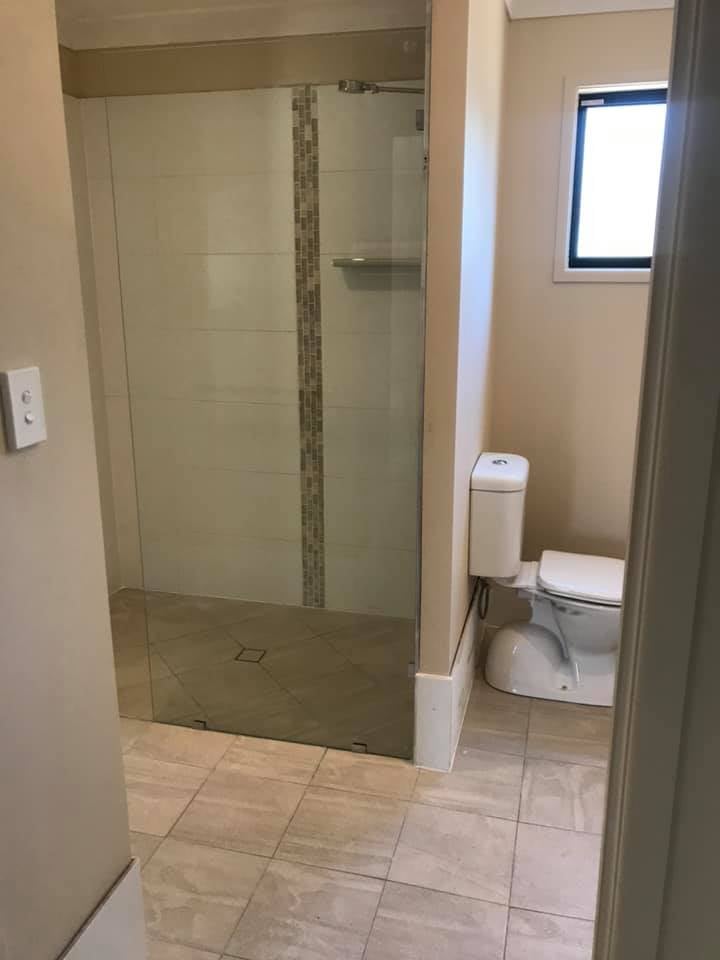Modern bathroom with tiled floor, glass-enclosed shower with vertical mosaic tile strip, and white toilet next to a small window.