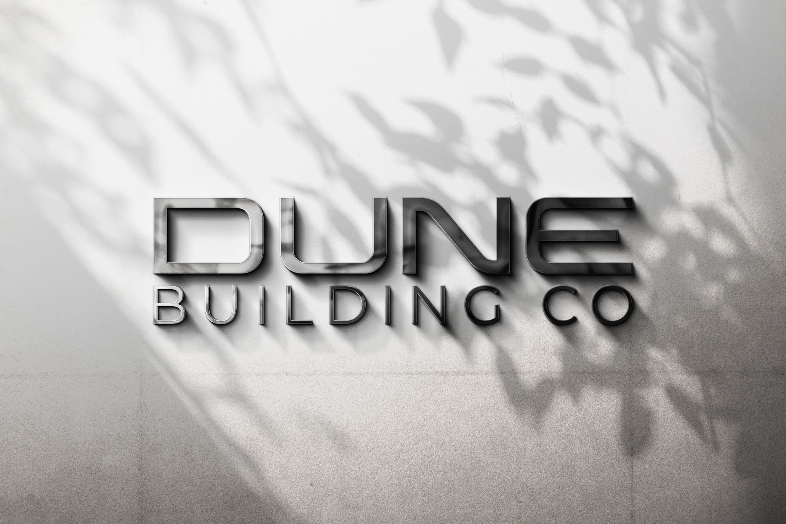 3D metallic sign reading 'DUNE BUILDING CO' on a light textured wall with shadow of leaves.