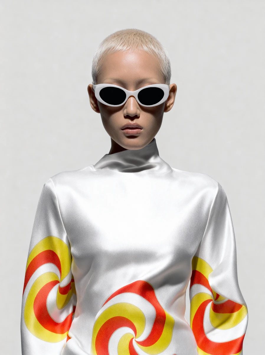 Person with short blond hair wearing white cat-eye sunglasses and a white satin top with red and yellow swirl patterns on the sleeves and torso.