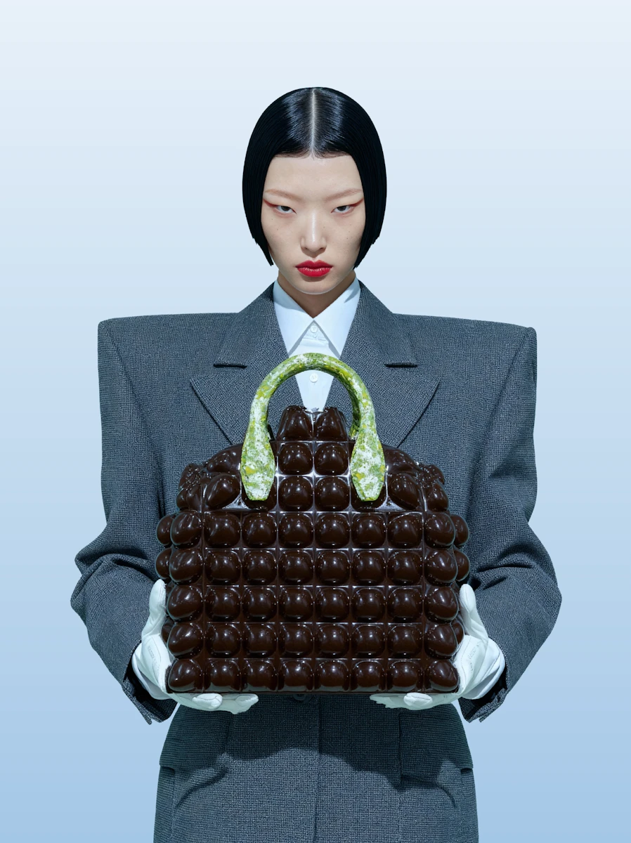 Model with sleek black hair and red lipstick wearing gray oversized blazer and white gloves holding a textured dark brown handbag with green handles.