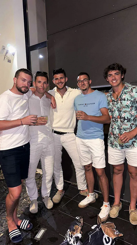 Five young men standing closely together indoors at night, smiling, two holding drinks, dressed casually in shorts, t-shirts, and summer clothing.