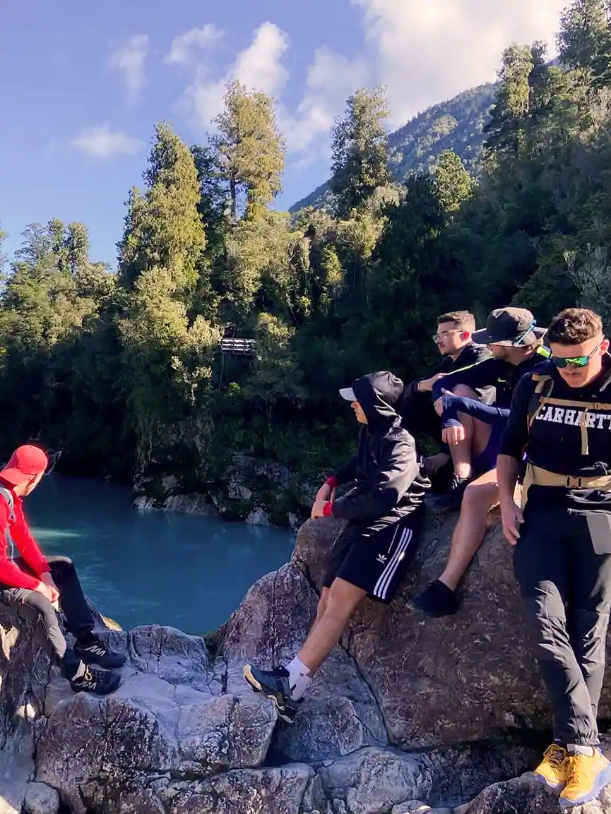 Group of young men sitting and standing on rocks near a turquoise river with forested hills in the background under a partly cloudy sky.