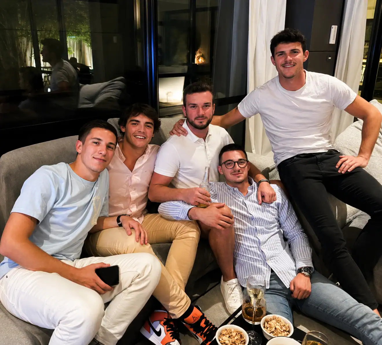 Five young men sitting closely on a gray couch in a modern living room with snacks and drinks on a table in front of them.