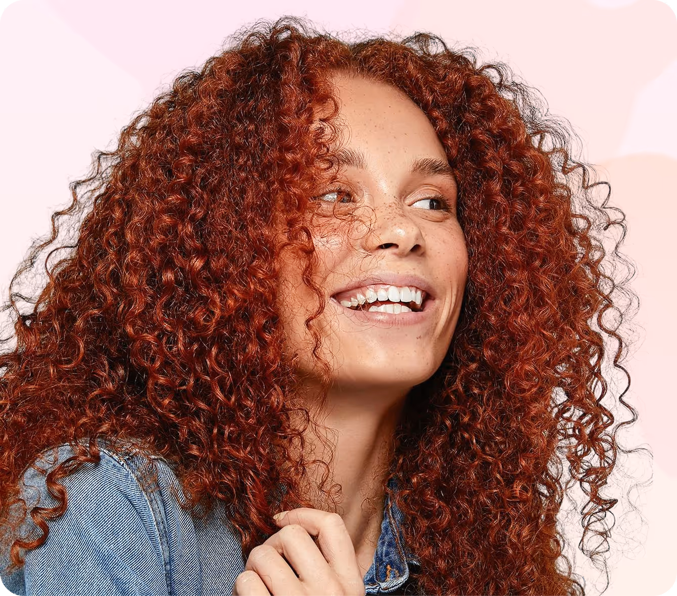 Smiling woman with curly red hair and freckles wearing a denim jacket against a light pink background.