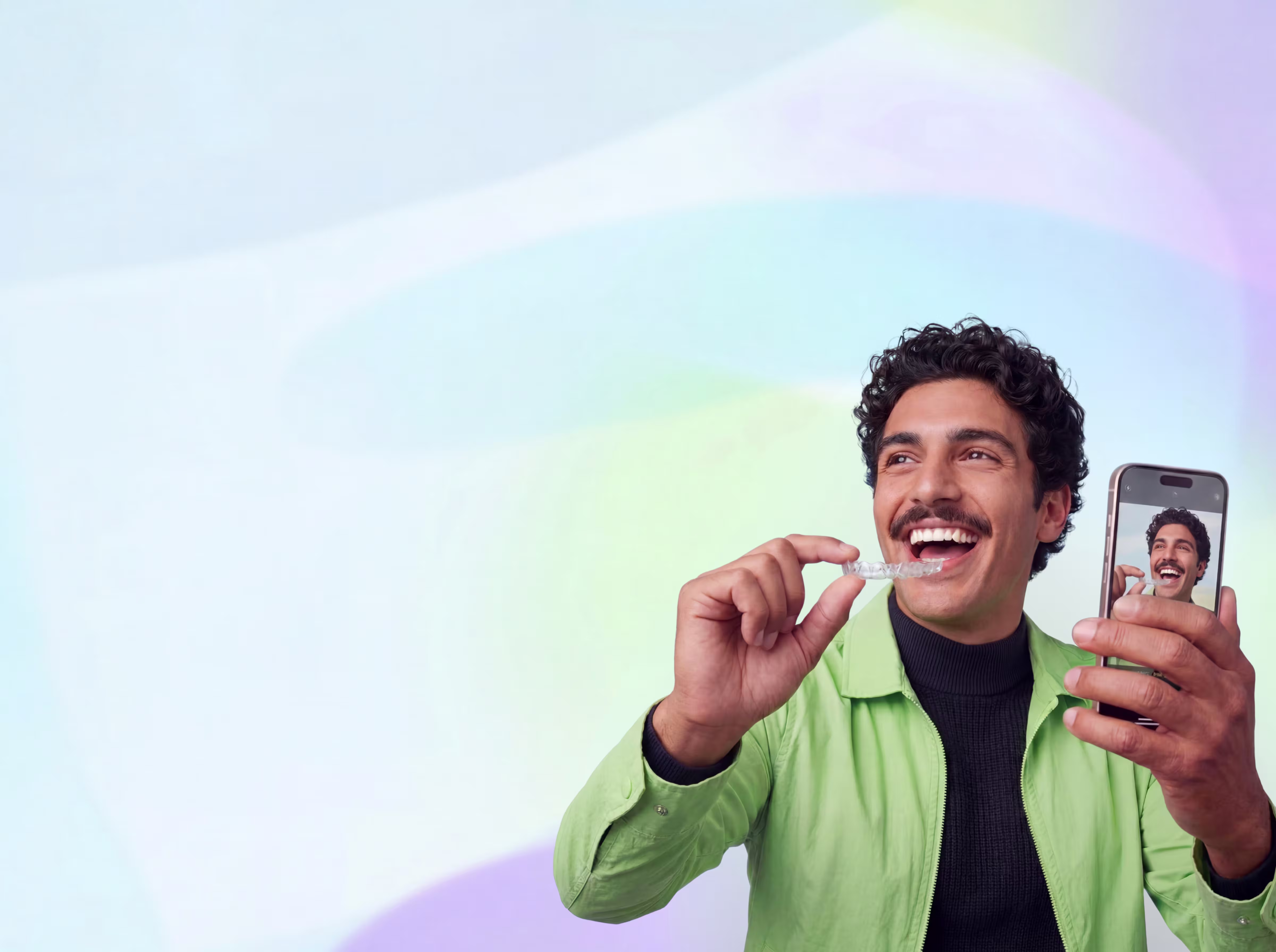 Smiling man holding a clear aligner near his mouth and showing a selfie on his smartphone.