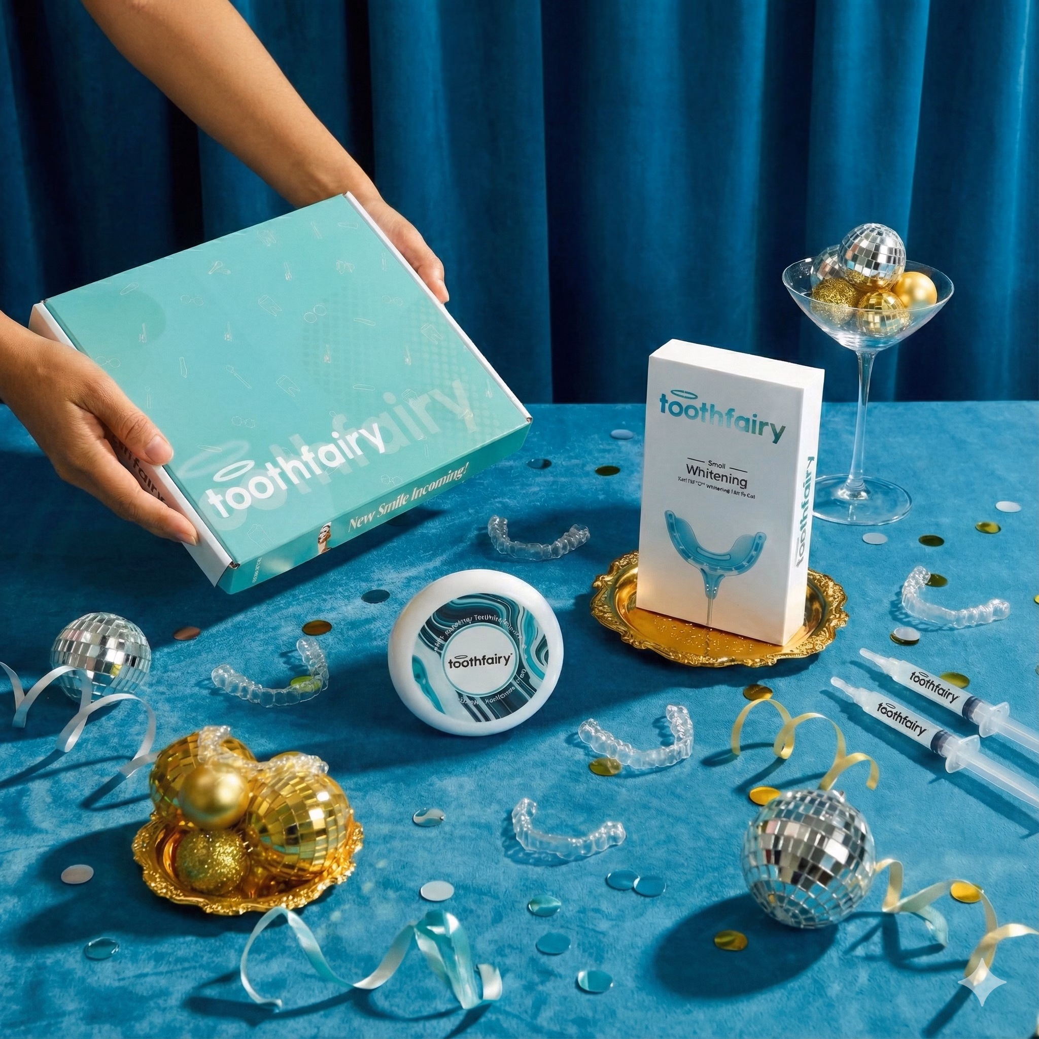 Hands holding teal Toothfairy box with whitening kit, trays, syringes, and decorative disco balls on blue table.