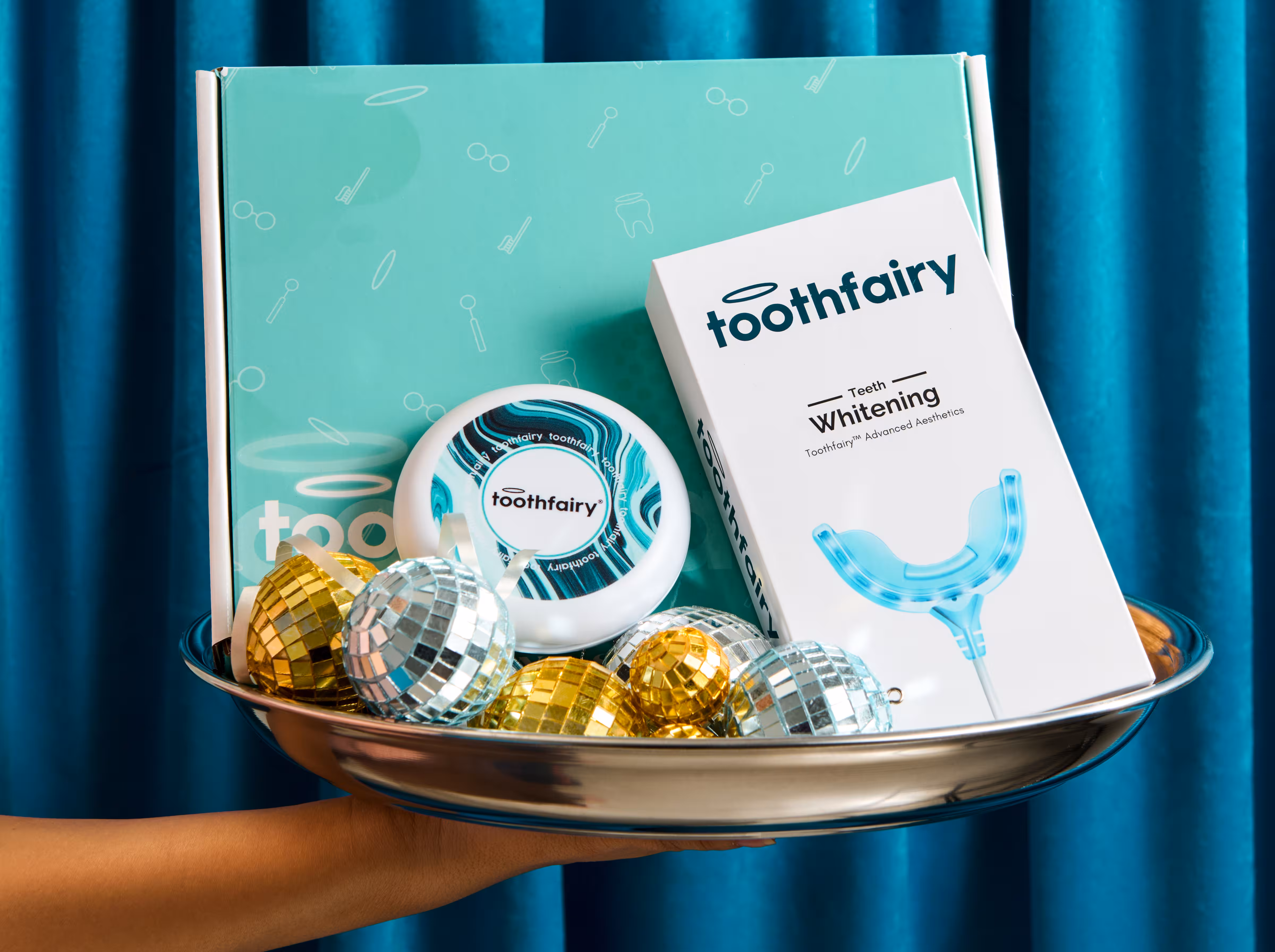 Hand holding silver tray with Toothfairy teeth whitening products and disco ball ornaments.