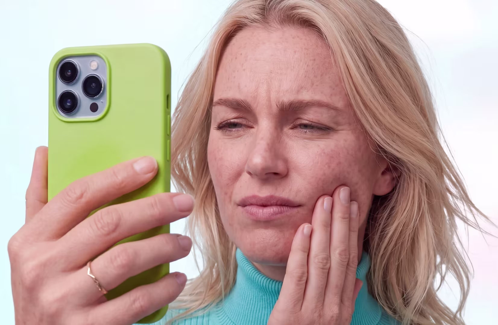 Woman holding green phone with worried expression touching her cheek as if feeling pain.
