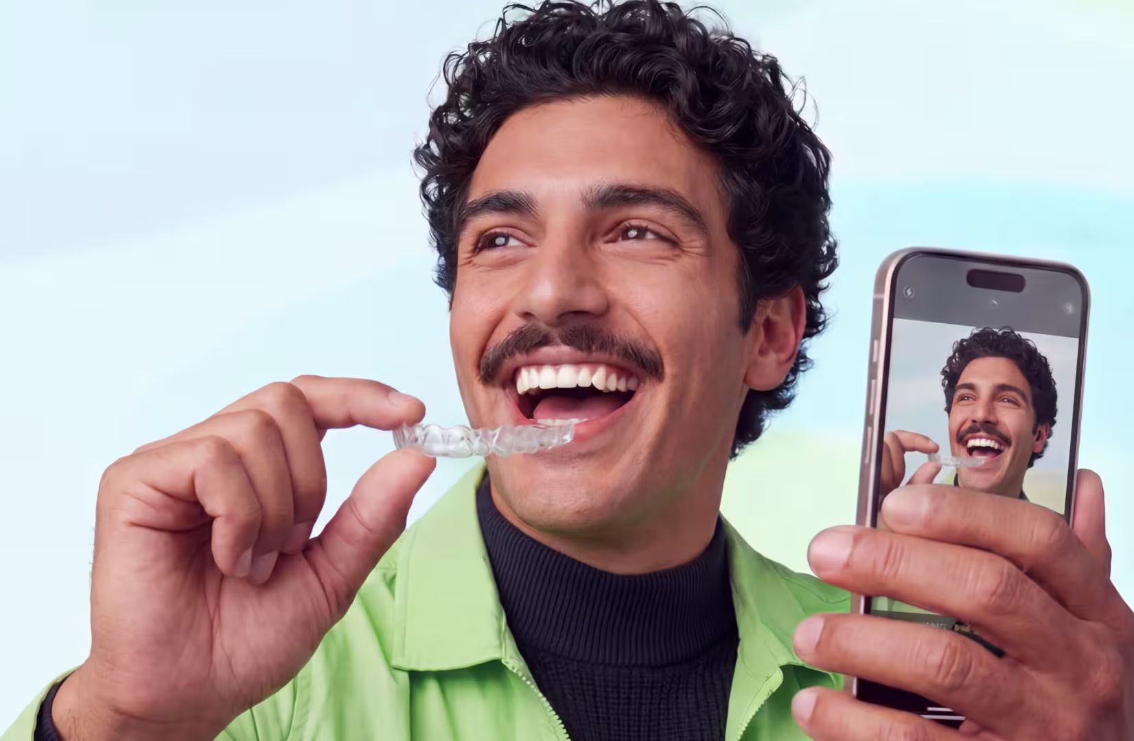 Man smiling while holding a clear dental aligner and a phone showing his image.