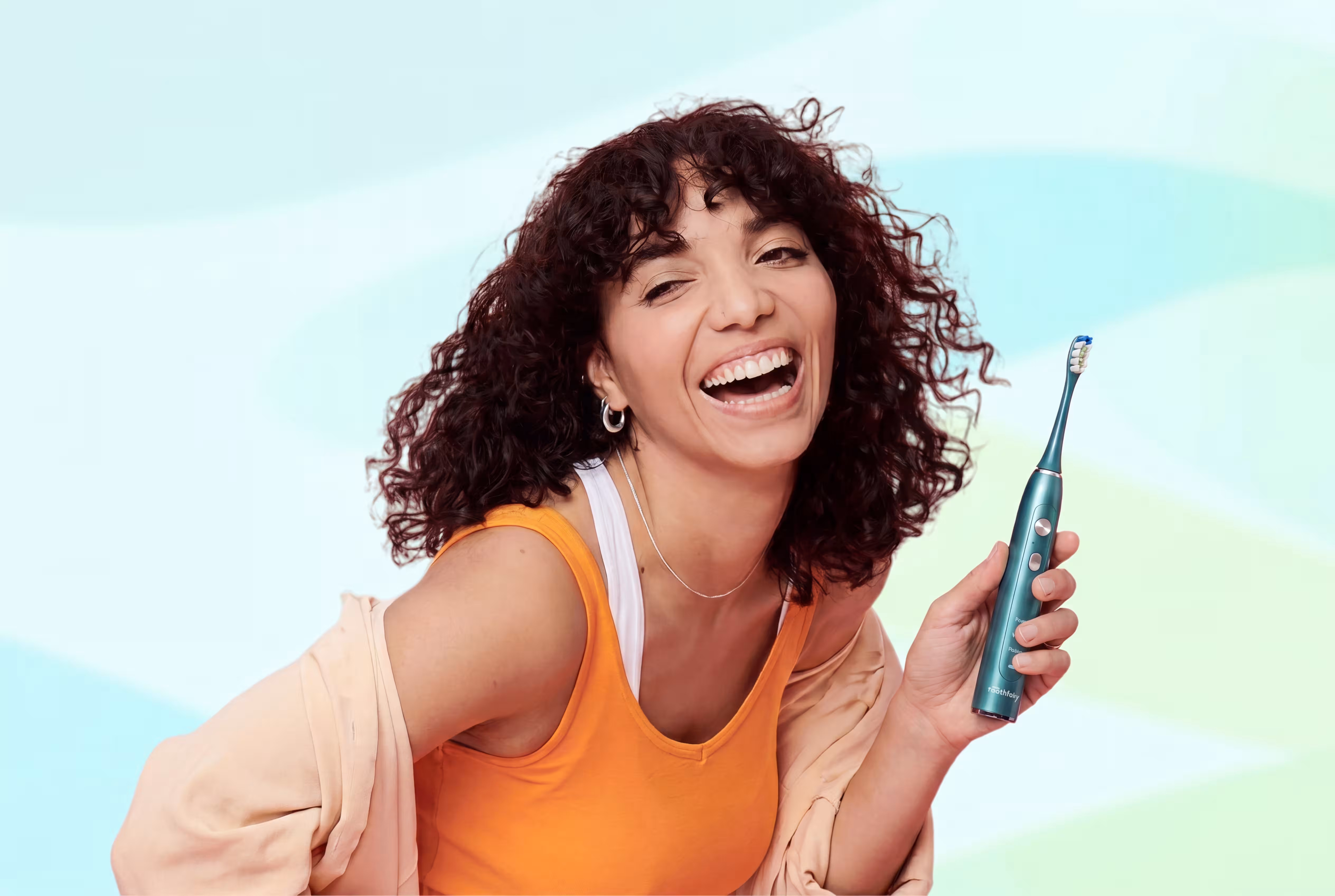 Smiling woman with curly hair holding a green electric toothbrush against a pastel background.