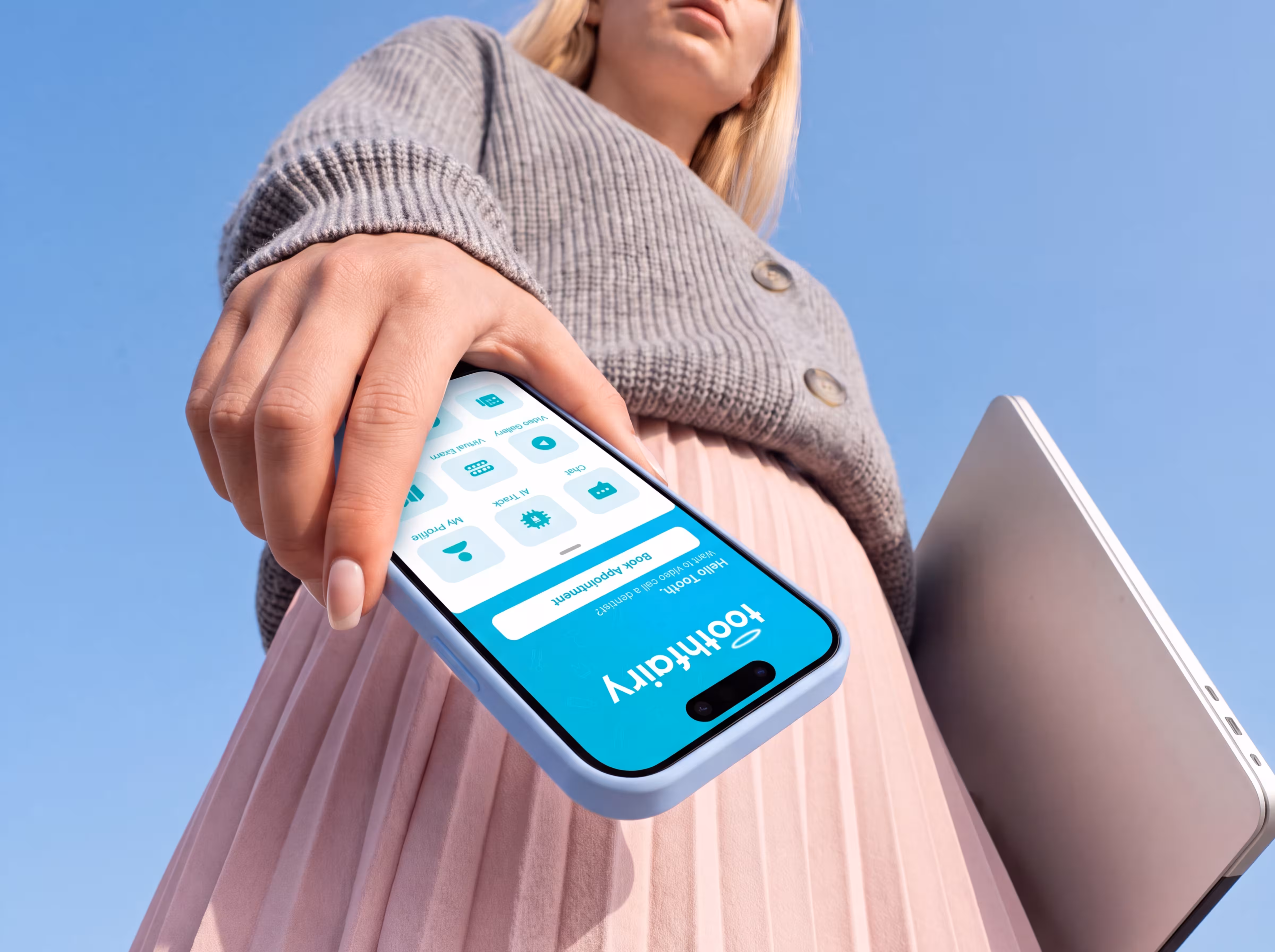 Woman holding smartphone showing Toothfairy app with icons and a laptop against blue sky.