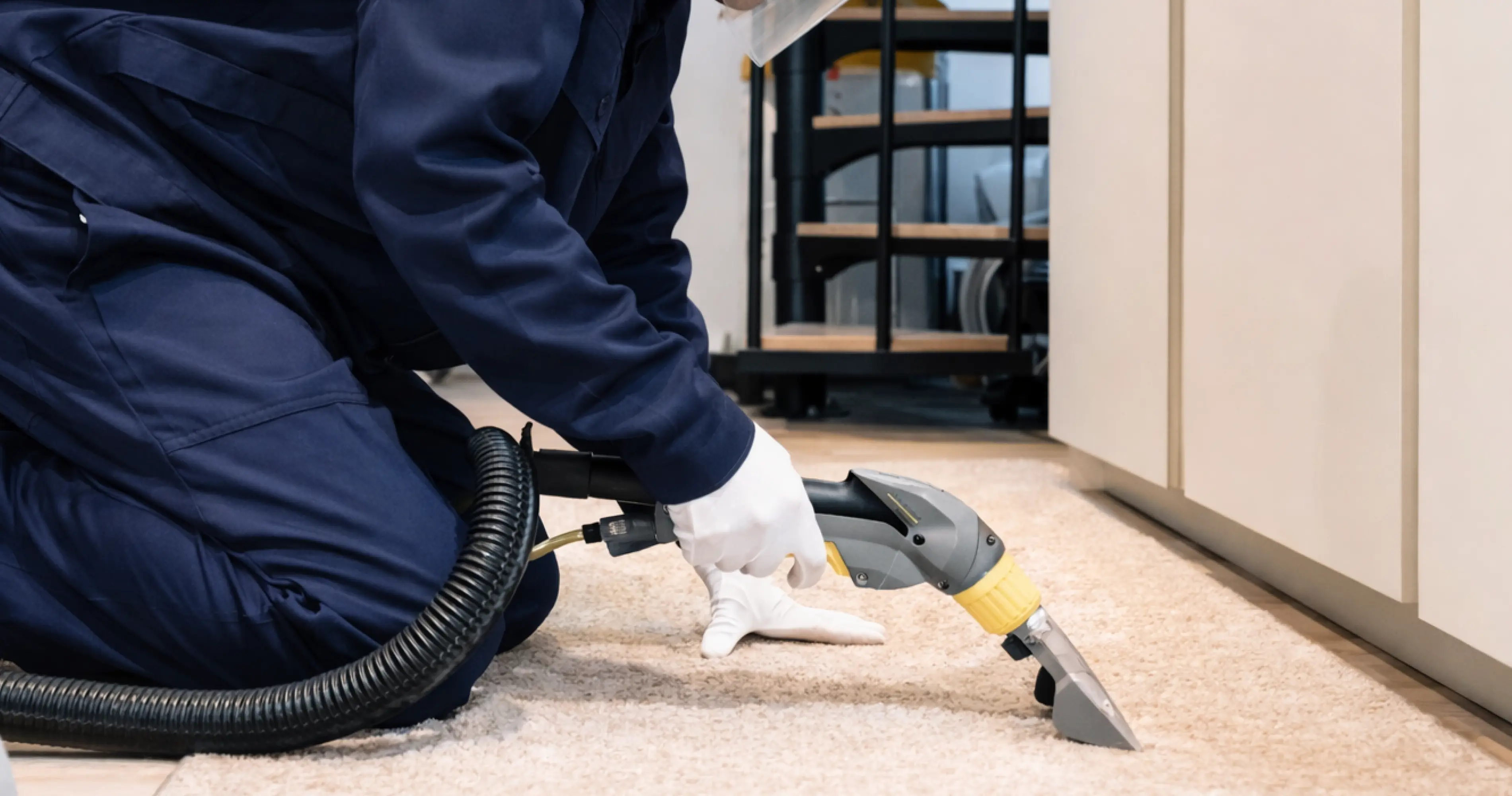 Professional cleaner in navy uniform and white gloves cleaning a beige carpet with a vacuum extraction tool near white cabinets.