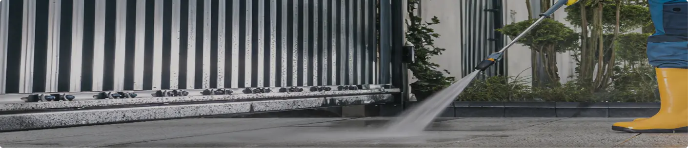 Person in yellow rubber boots pressure washing a tiled outdoor surface near a metal gate and garden.