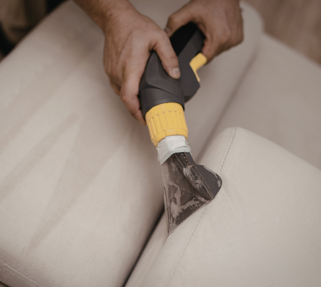 Person using a steam or suction cleaning tool on a beige upholstered sofa.