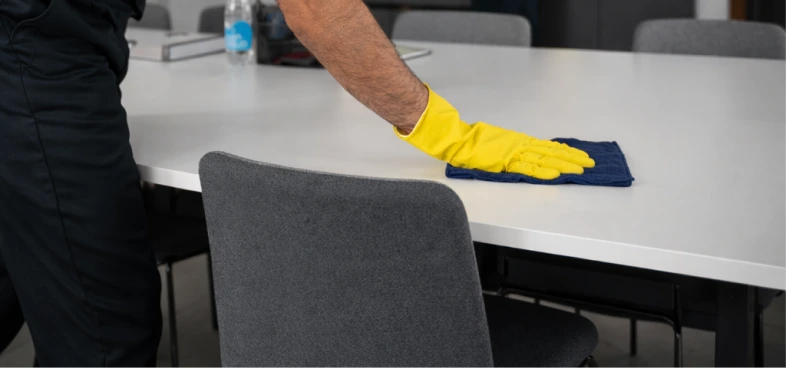 Person wearing yellow gloves wiping a white table with a blue cloth in an office setting.