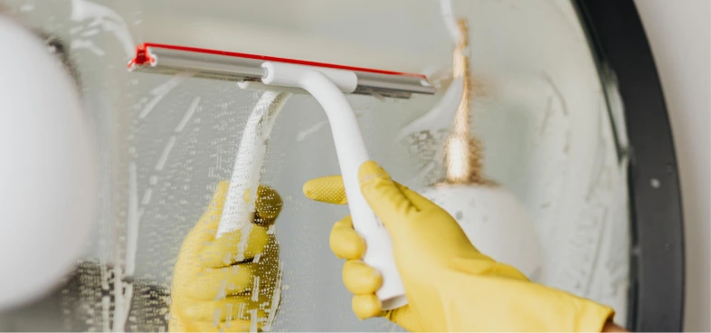 Hand wearing yellow gloves wiping a soapy glass surface with a white and red squeegee.