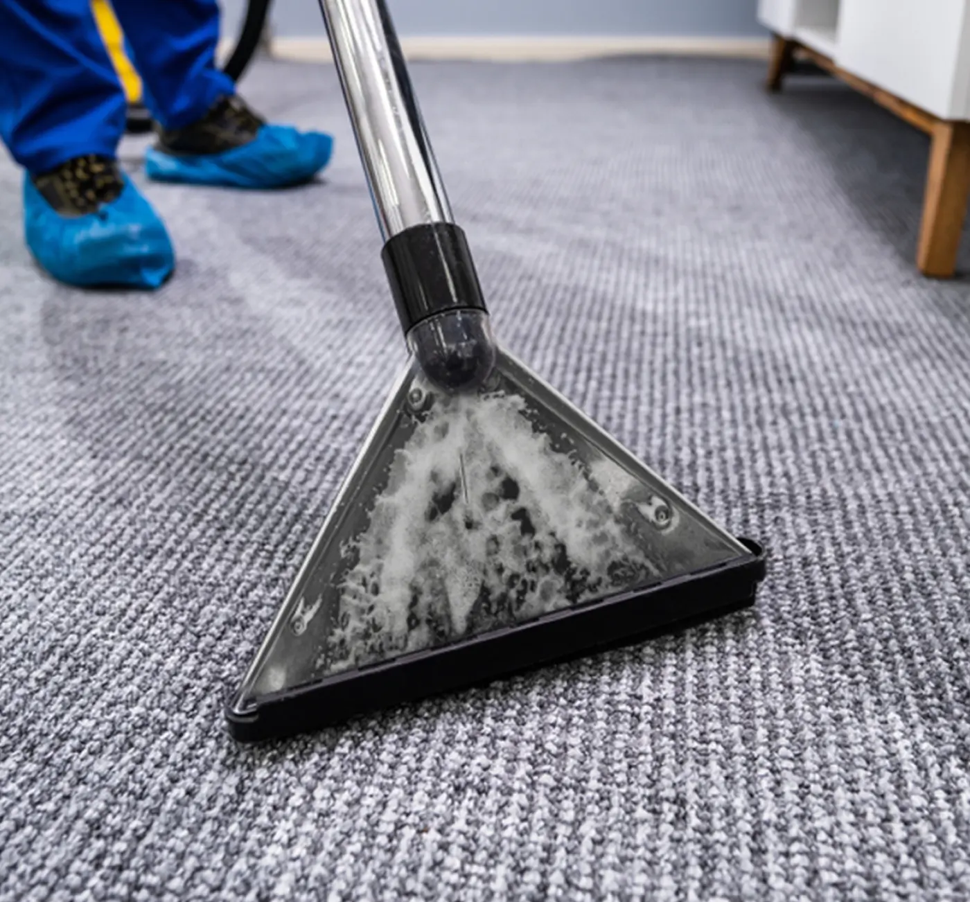 Close-up of a carpet cleaning machine with foam inside its transparent nozzle on a gray textured carpet.