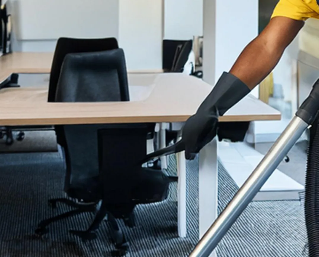 Person wearing black gloves vacuuming carpet under a desk in an office with black chairs.