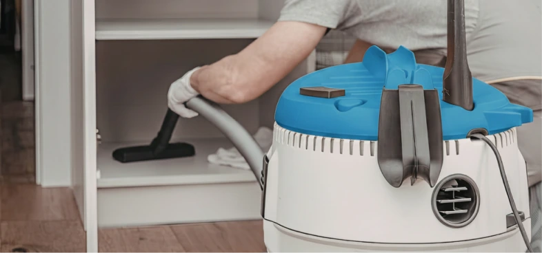 Person vacuuming inside a white cabinet with a canister vacuum cleaner featuring a blue top and black attachments.