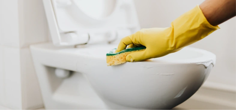 Hand in a yellow glove cleaning the rim of a white toilet bowl with a sponge.