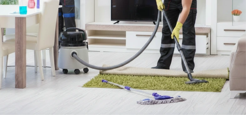 Person using a vacuum cleaner to clean a green rug in a modern living room with a mop on the floor nearby.