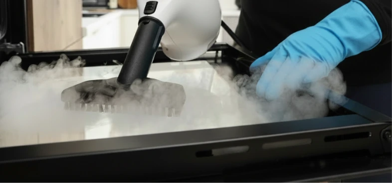 Person wearing blue glove using a steam cleaner to clean an oven interior with steam visible.