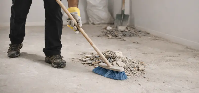 Person wearing gloves sweeping debris and rubble from a concrete floor with a broom.