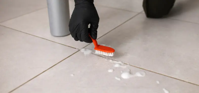 Hand wearing a black glove scrubbing tile floor grout with an orange brush and cleaning foam.