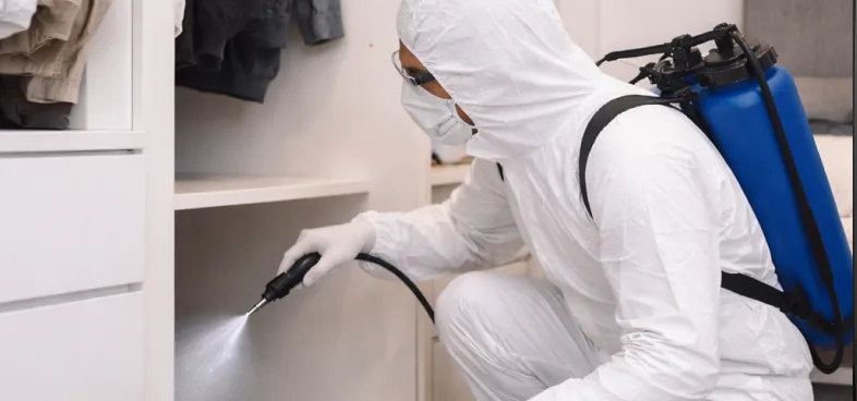 Person in white protective suit spraying disinfectant inside a cabinet with a blue backpack sprayer.