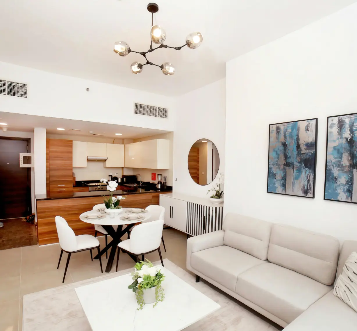 Modern open-plan living room and kitchen with beige sofa, round dining table with white chairs, and abstract blue paintings on wall.