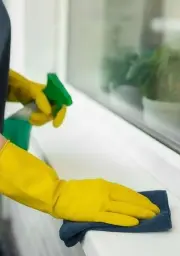 Person wearing yellow gloves cleaning a window sill with a blue cloth and green spray bottle.