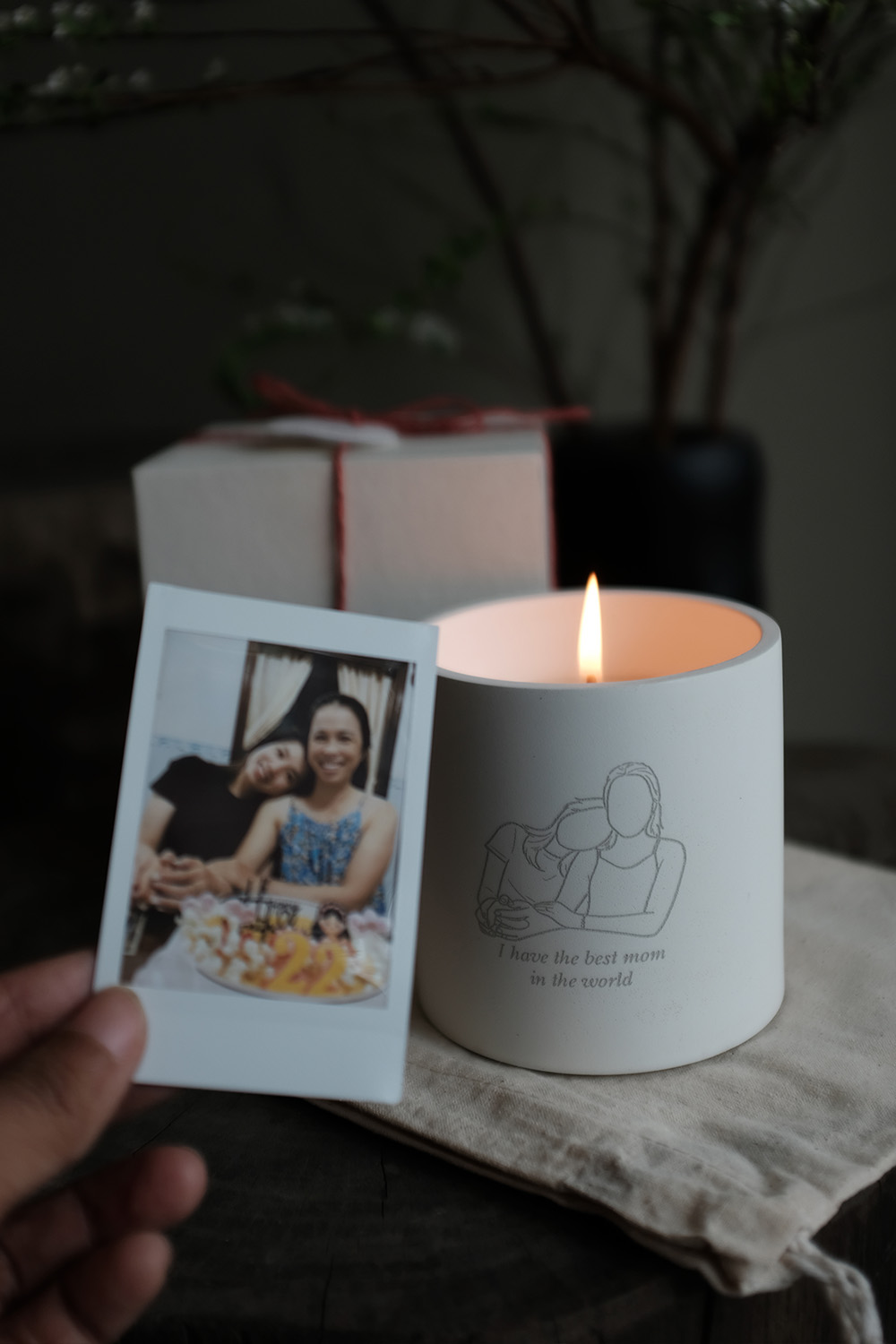 Lit white candle with a line art of two women hugging and text saying 'I have the best mom in the world,' next to a hand holding a Polaroid photo of two smiling women at a birthday with a cake.