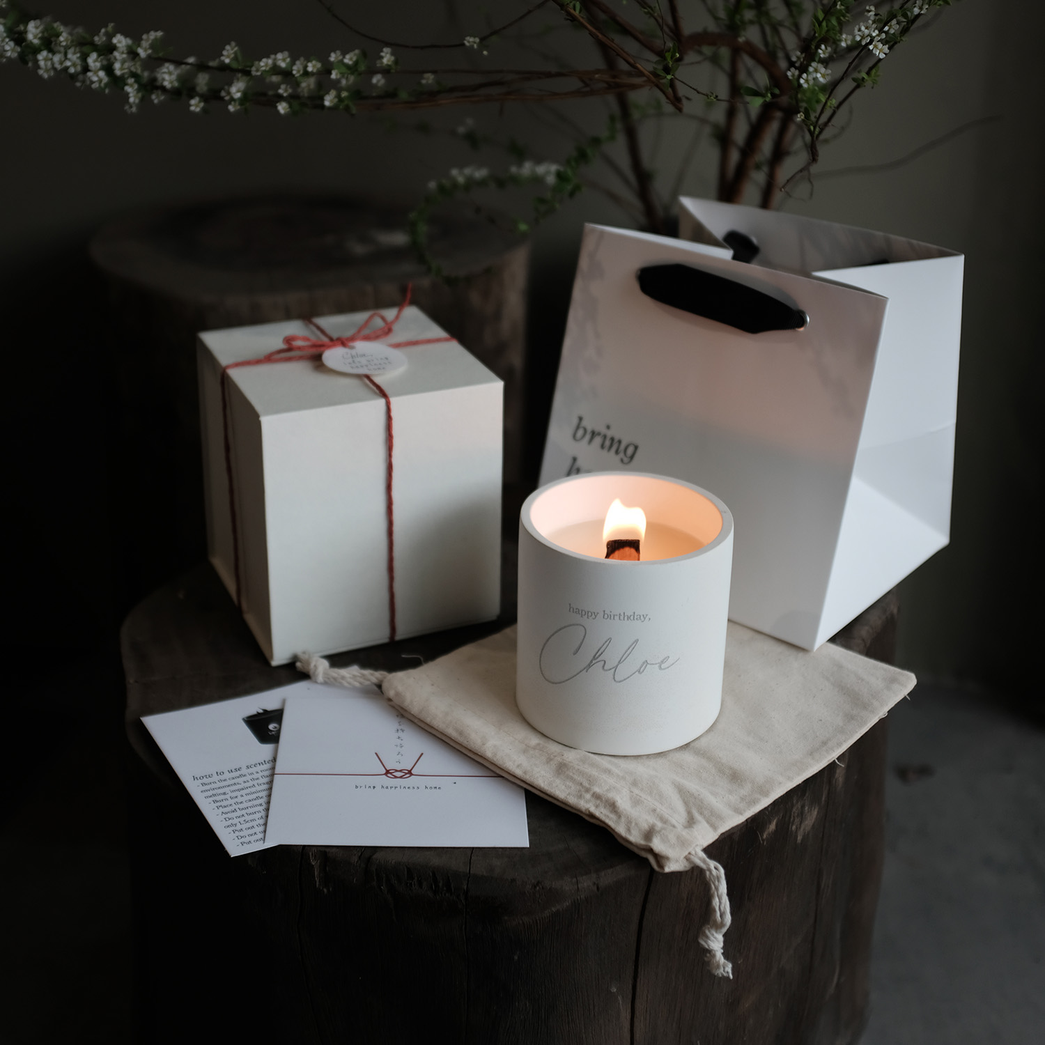 Lit birthday candle in a white holder with the name Chloe, next to a gift box, a white gift bag, and cards on a wooden surface.
