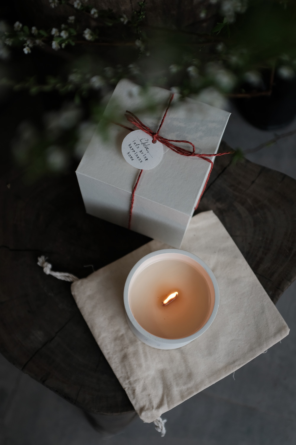 Lit candle in a white container placed on a beige cloth next to a white gift box tied with red string and a round tag.