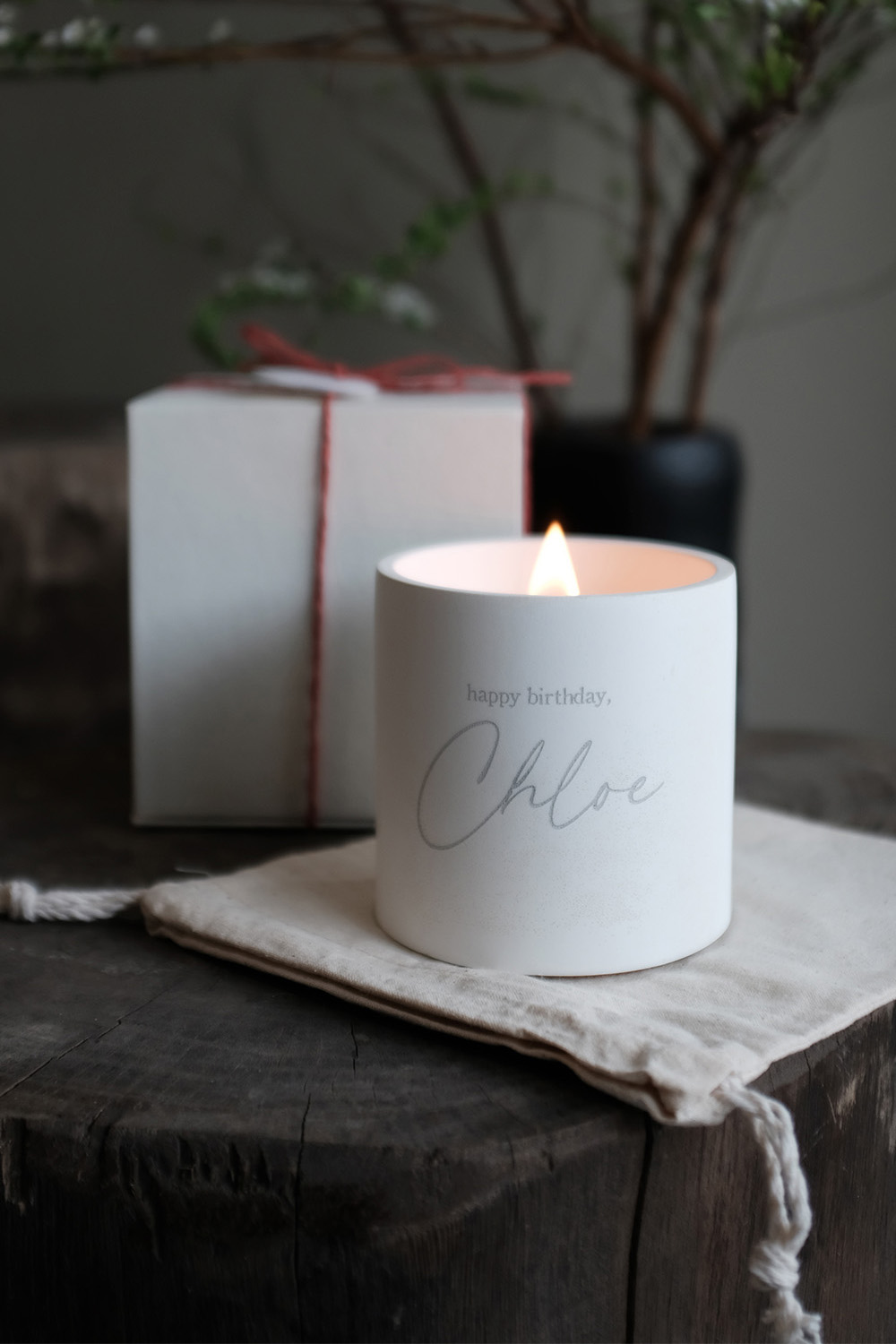 Lit white candle with 'happy birthday, Chloe' inscription placed on a cloth on a wooden surface, with a gift box blurred in the background.