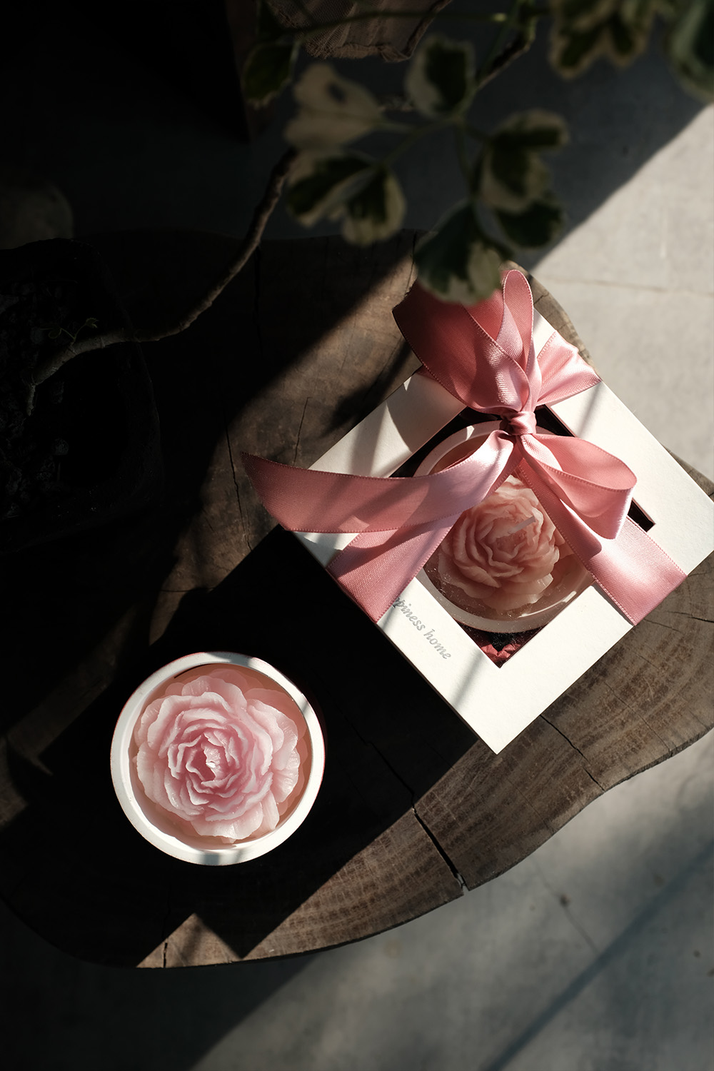 Pink flower-shaped candle in a white container alongside a similar candle in a white gift box with a pink ribbon, placed on a wooden surface.