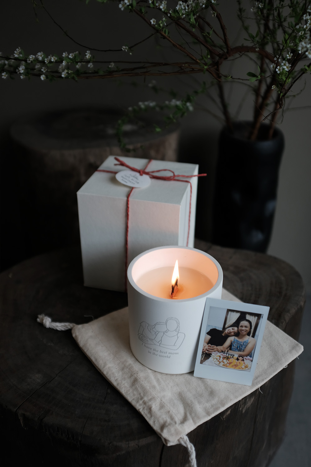 Lit candle in white holder with sketch and text 'I have the best mom in the world,' white gift box tied with red string, and a Polaroid photo of two smiling women on rustic wooden surface.