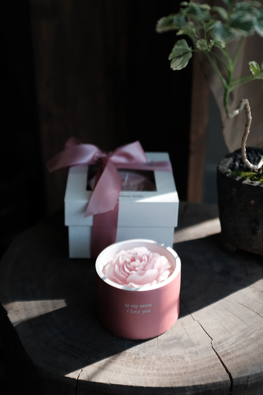 Pink flower-shaped candle in a pink container with the words 'to my mom i love you' on a wooden surface next to a white gift box tied with a pink ribbon.