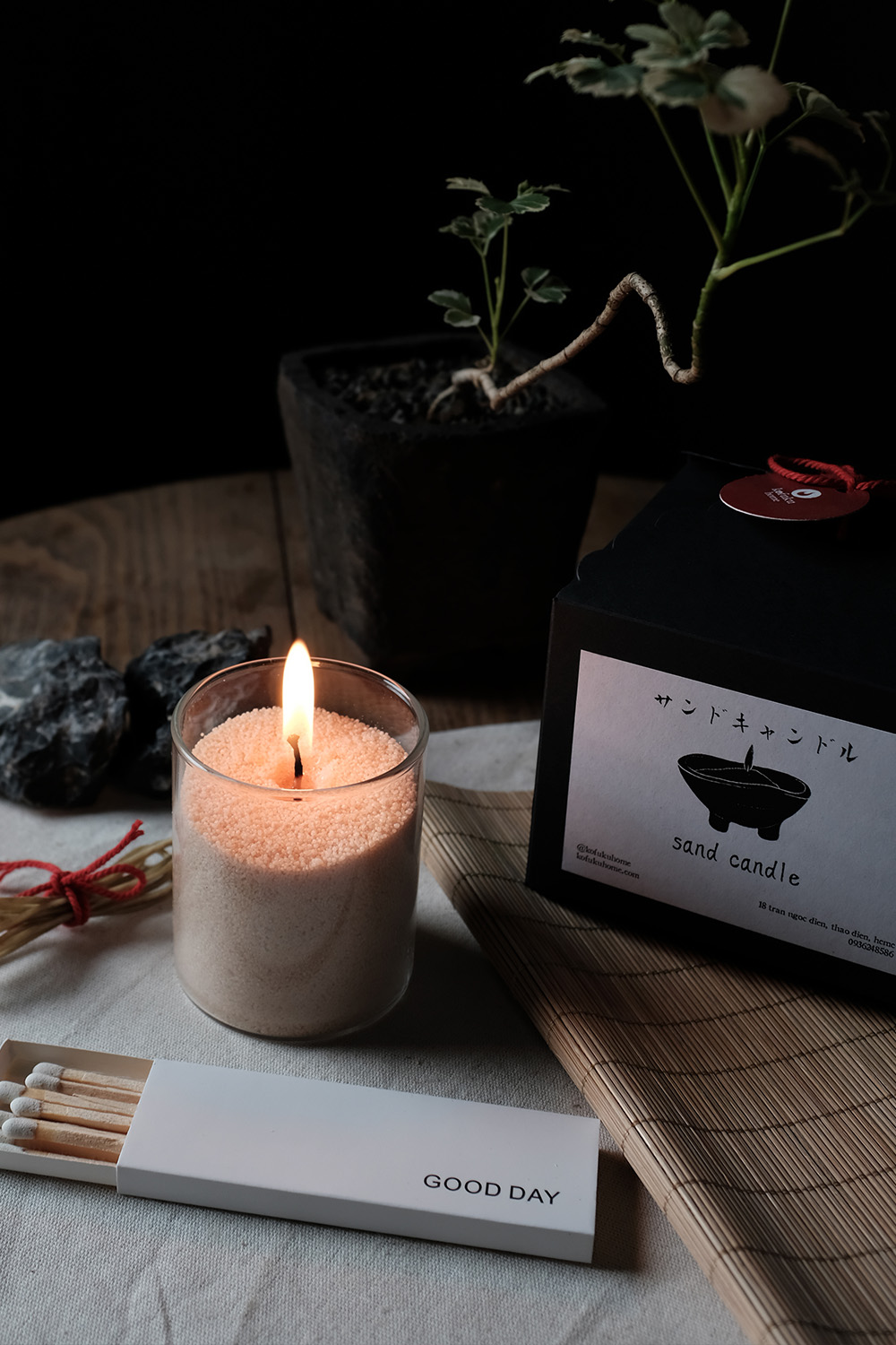 Burning sand candle in a glass container next to a black box labeled 'sand candle,' a plant in a black pot, and an open matchbox with 'GOOD DAY' text.