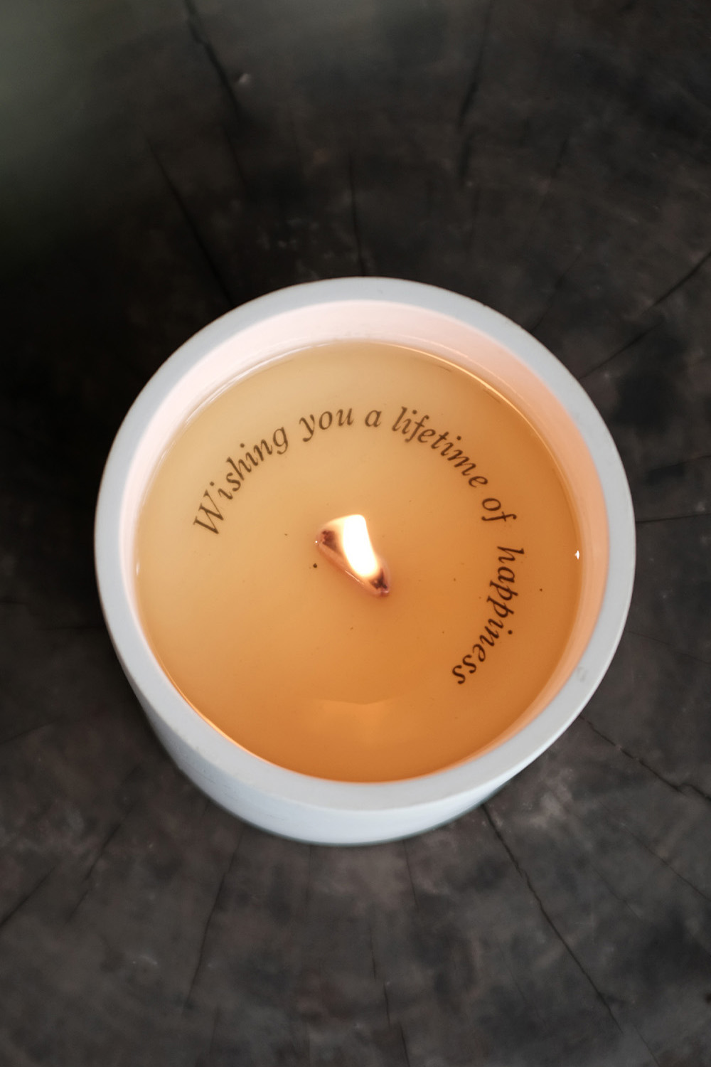 Lit candle in a white container with a message around the flame saying 'Wishing you a lifetime of happiness.'
