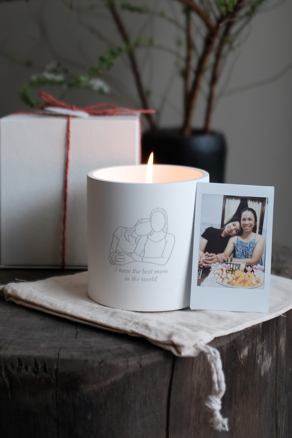 Lit white candle with line art of two people and text 'I have the best mom in the world', a photo of two smiling women, and a gift box tied with red string on a wooden surface.